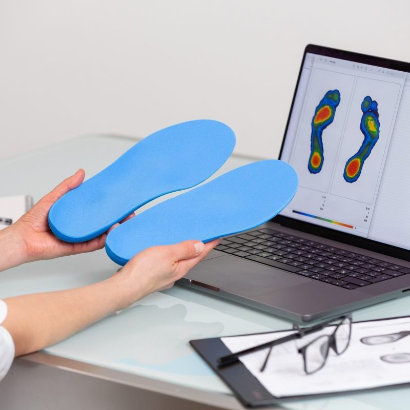 Female doctor orthopedist holding custom made insoles in a clinic in front of the laptop with a test feet picture.