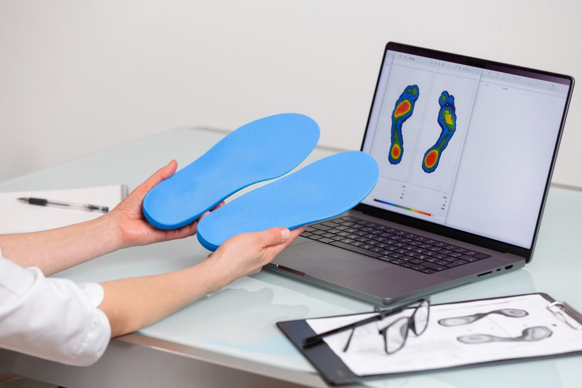 Female doctor orthopedist holding custom made insoles in a clinic in front of the laptop with a test feet picture.